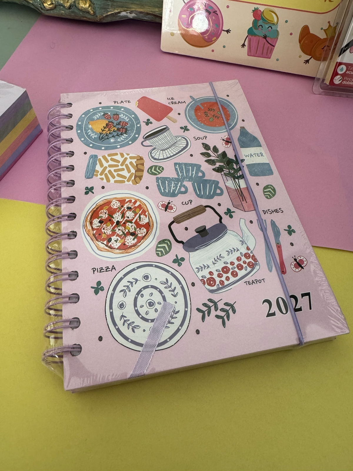 Agenda Brena 2027 de espiral en color rosa con ilustraciones de comida y cocina sobre fondo rosa y amarillo