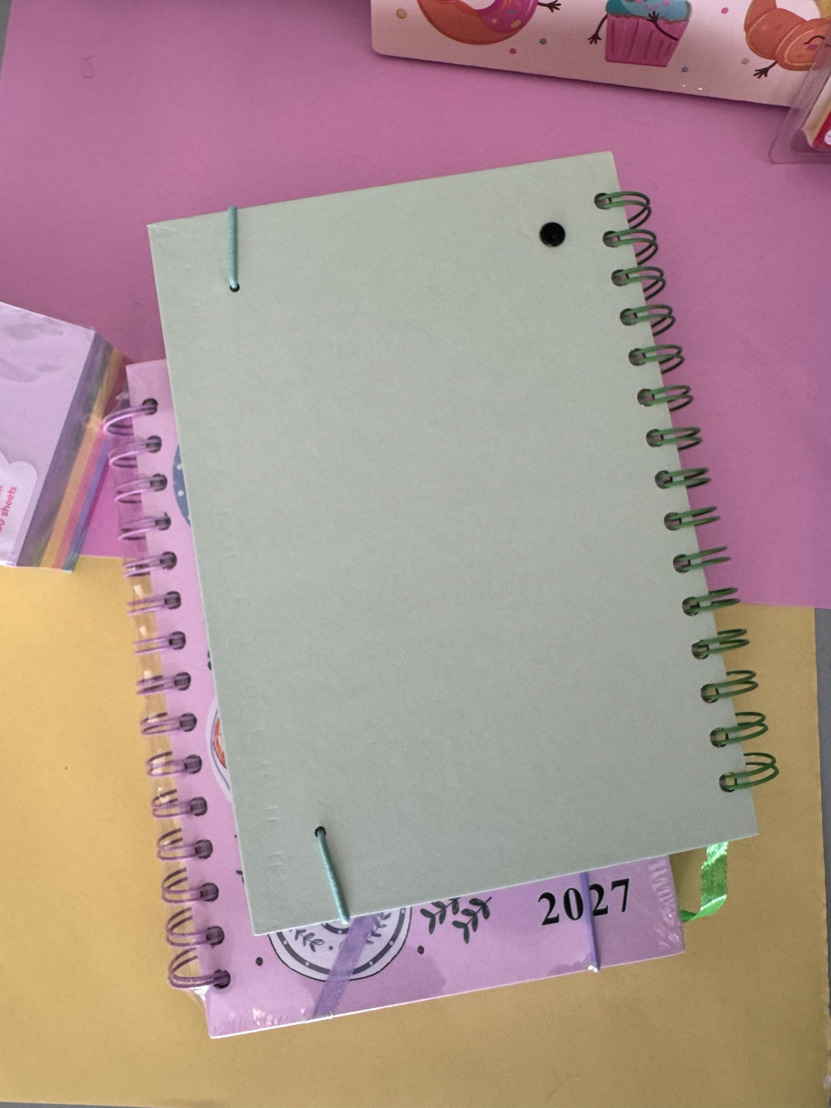 Agenda Brena 2027 con espiral metálica, cierre elástico y cubierta verde suave sobre fondo de papelería colorida