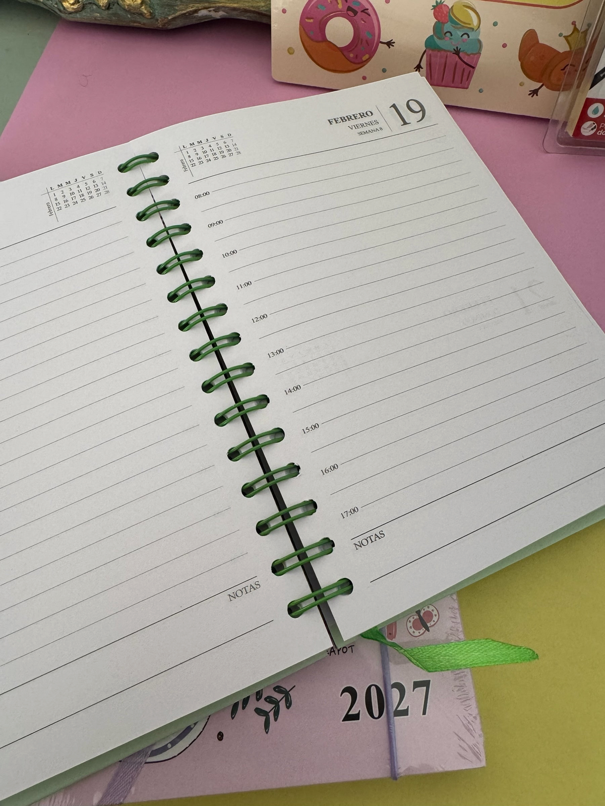 Agenda Brena 2027 de espiral con vista diaria detallada y espacio para notas, sobre fondo colorido con ilustraciones.