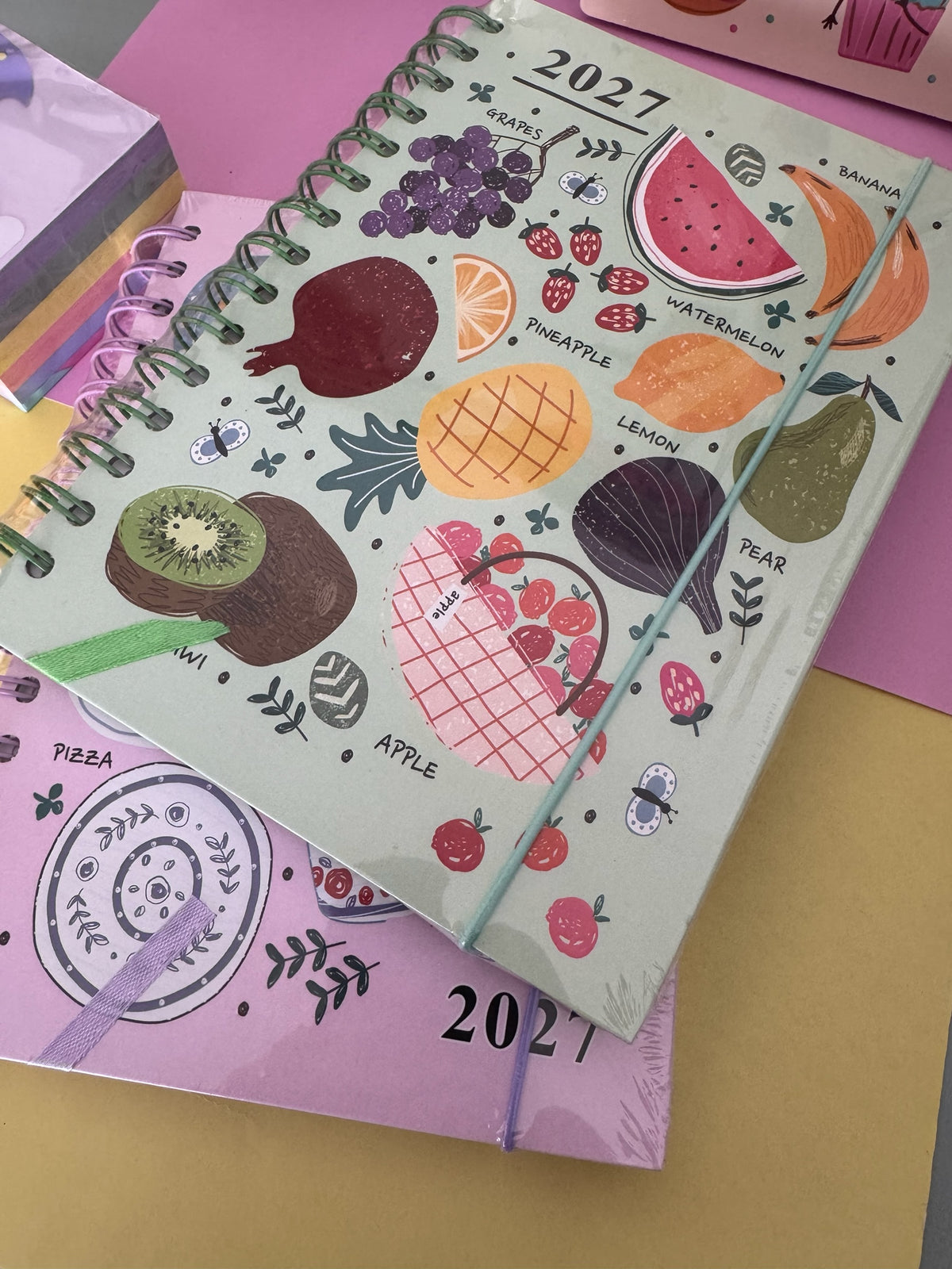 Agenda semanal Brena 2027 con estampado de frutas coloridas sobre fondo verde, espiral metálica y cierre elástico.