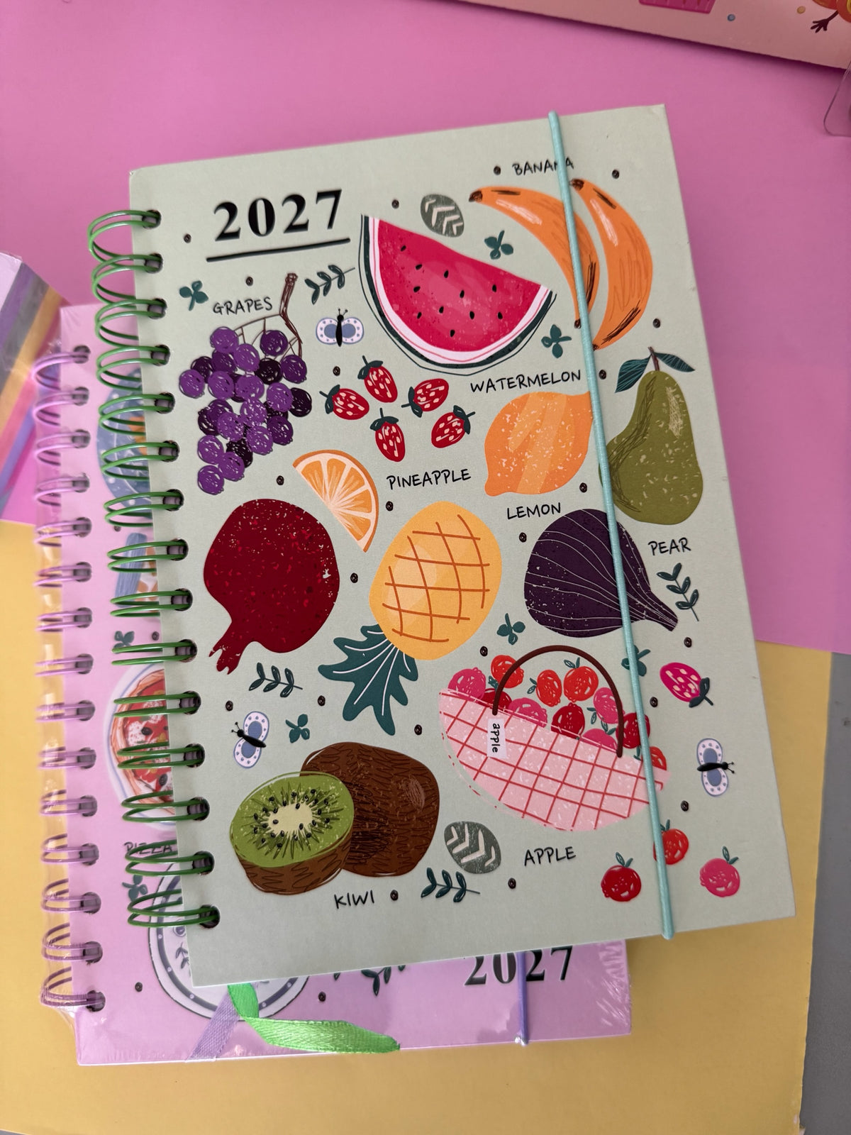 Agenda semanal Brena 2027 con diseño de frutas coloridas sobre fondo verde, encuadernación en espiral y cierre elástico