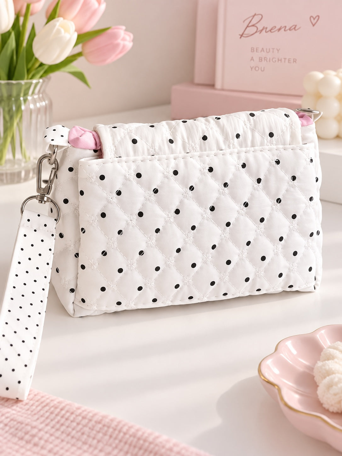 Bolso bandolera acolchado Brena blanco con lunares negros, correa ajustable y detalles en rosa sobre mesa blanca.