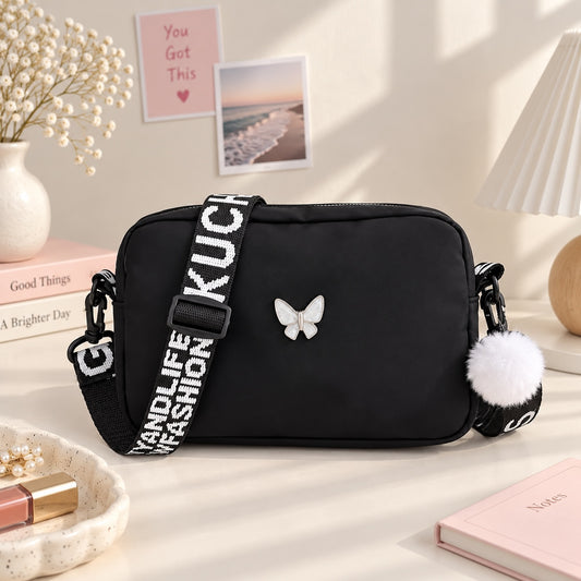 Bolso bandolera Brena negro con detalle de mariposa, correa tipográfica ajustable y pompón decorativo blanco.