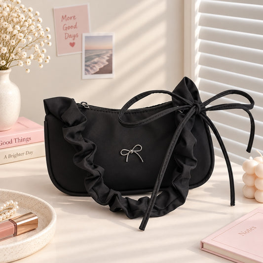 Bolso de hombro Brena negro tipo baguette con volantes decorativos y lazo metálico en un entorno elegante