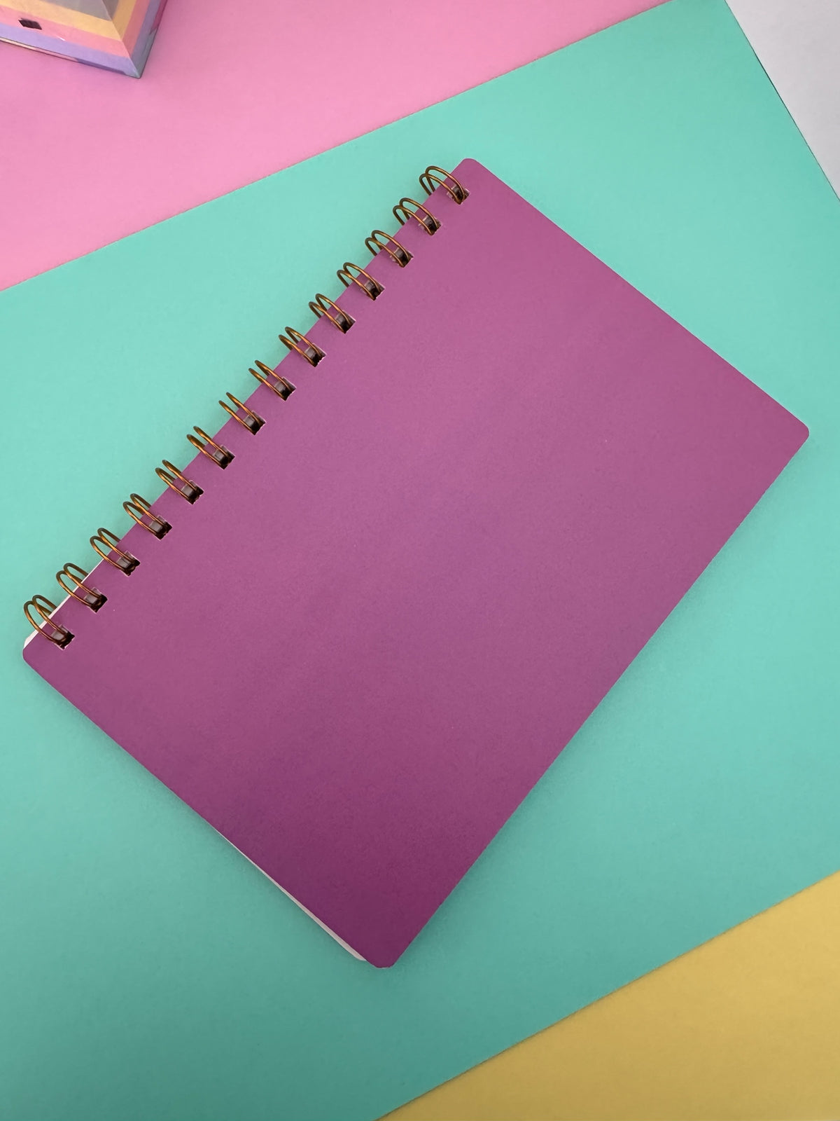 Planner semanal Brena con espiral metálica dorada y portada de color morado sobre fondo de tonos pastel